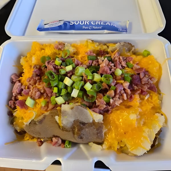 A loaded baked potato with cheese, bacon bits, and green onions in a takeout container with sour cream.