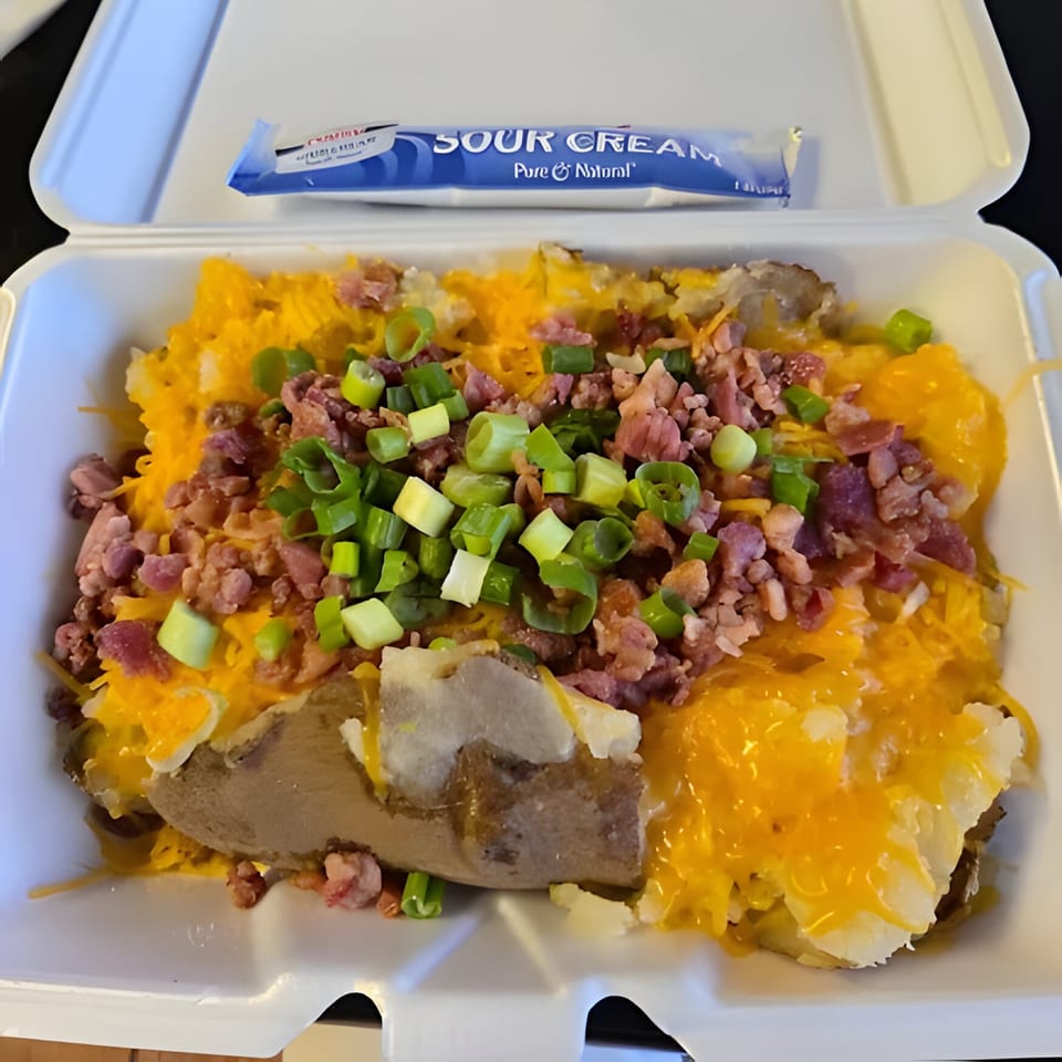 A loaded baked potato with cheese, bacon bits, and green onions in a takeout container with sour cream.